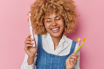 Positive woman with curly hair holds different kinds of toothbrushes takes care of oral hygiene smiles broadly shows white perfect teeth wears white shirt and denim sarafan isolated on pink background