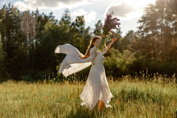 Young beautiful woman, wearing white dress, holding flowers and dancing on the meadow. Girl joying...