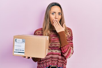 Beautiful hispanic woman holding delivery package covering mouth with hand, shocked and afraid for...