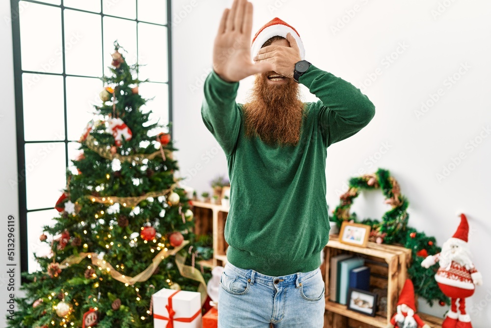 Poster redhead man with long beard wearing christmas hat by christmas tree covering eyes with hands and doi