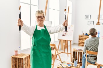 Two senior paint student smiling happy painting at art studio. Woman standing with smile on face holding paintbrushes.