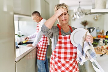 Middle age caucasian couple wearing apron washing dishes at home stressed and frustrated with hand on head, surprised and angry face