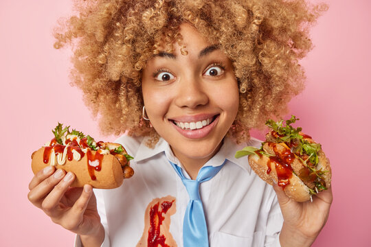 Close Up Shot Of Cheerful Woman Looks With Big Surprised At Camera Smiles Broadly Holds Fast Food Enjoys Eating Tasty Sandwich And Hot Dog Dressed In Formal Clothes Smeared As Being Careless