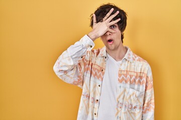 Young man wearing casual summer shirt peeking in shock covering face and eyes with hand, looking through fingers with embarrassed expression.