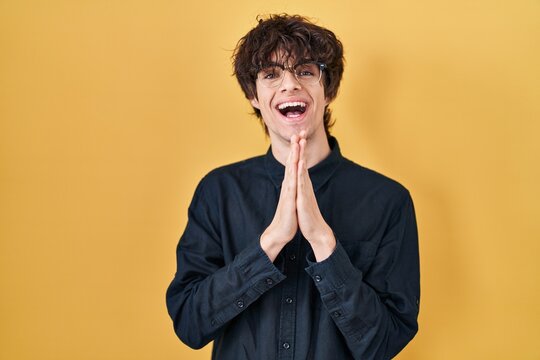 Young man wearing glasses over yellow background praying with hands together asking for forgiveness smiling confident.