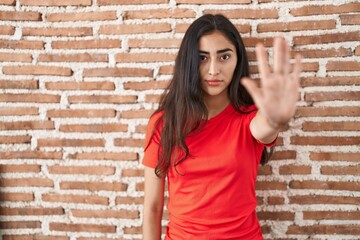Young teenager girl standing over bricks wall doing stop sing with palm of the hand. warning expression with negative and serious gesture on the face.