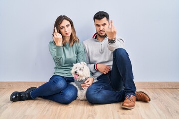 Obraz na płótnie Canvas Young hispanic couple sitting on the floor with dog showing middle finger, impolite and rude fuck off expression