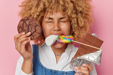 Indoor shot of positive woman with curly hair enjoys eating delicious sweet treats holds cookie bar...