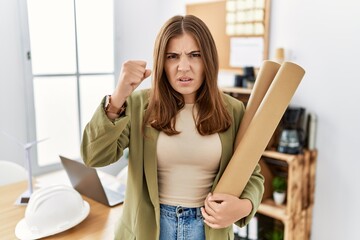Fototapeta na wymiar Young brunette woman holding paper blueprints at the office annoyed and frustrated shouting with anger, yelling crazy with anger and hand raised
