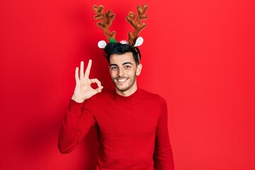 Young hispanic man wearing cute christmas reindeer horns smiling positive doing ok sign with hand...