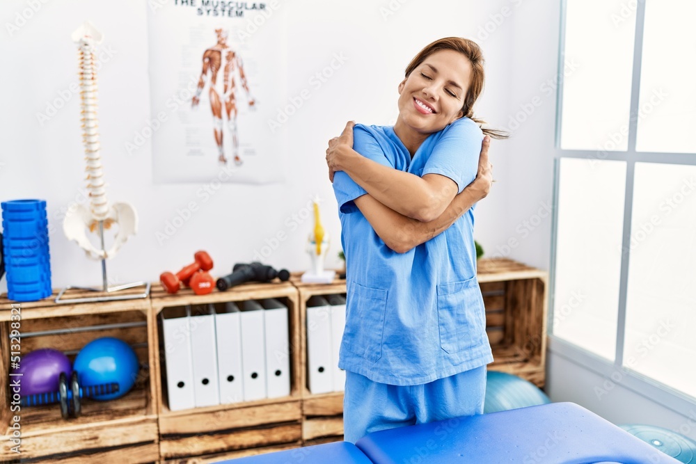 Poster Middle age hispanic physiotherapist woman working at pain recovery clinic hugging oneself happy and positive, smiling confident. self love and self care