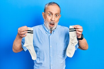 Handsome senior man with beard holding clean andy dirty socks in shock face, looking skeptical and...