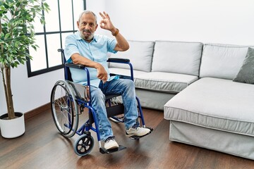 Handsome senior man sitting on wheelchair at the living room smiling positive doing ok sign with hand and fingers. successful expression.