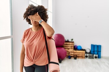 African american woman with afro hair holding yoga mat at pilates room covering eyes with hand,...