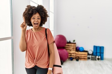 Fototapeta na wymiar African american woman with afro hair holding yoga mat at pilates room pointing finger up with successful idea. exited and happy. number one.
