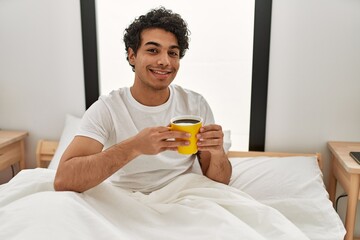 Young hispanic man drinking coffee sitting on the bed at bedroom.