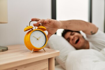 Young hispanic man turning off alarm clock lying on the bed at bedroom.