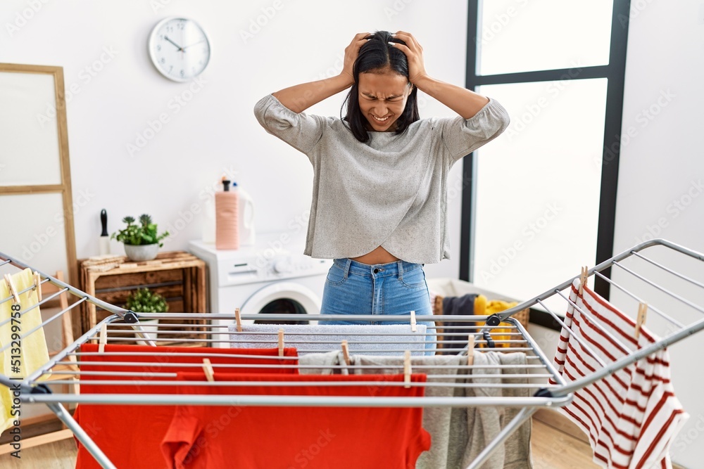 Sticker young hispanic woman putting fresh laundry on clothesline suffering from headache desperate and stre