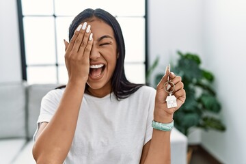 Young hispanic woman holding keys of new home covering one eye with hand, confident smile on face and surprise emotion.