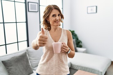 Middle age woman drinking a cup coffee at home approving doing positive gesture with hand, thumbs...