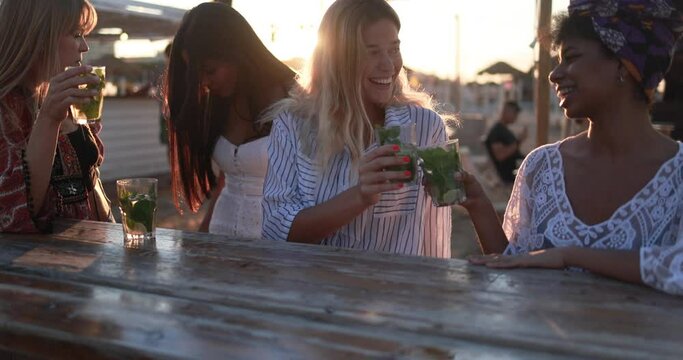 Happy multiracial girls having fun cheering with mojitos at beach party with sunset in background