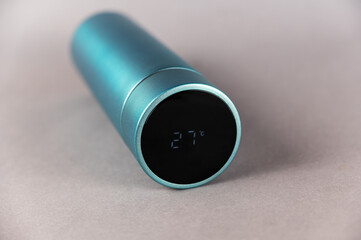 Modern thermos lies on a gray background. Black lid with LED digital display. Temperature reading 27 degrees Celsius. Cylindrical vacuum flask of turquoise color. Container for hot and cold drinks.