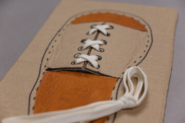 Card for tying laces is against gray background. White shoelace is inserted into the eyelets. The second shoelace lies next to it. Homemade card made of three-ply brown cardstock. Series part.