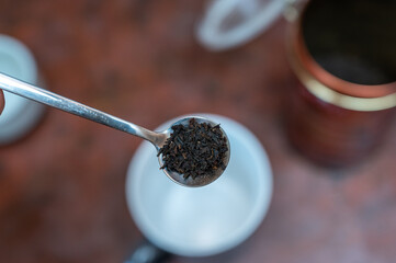 Middle-aged man holding spoonful of dry tea. Crushed black tea leaves in spoon over red table. Cup...