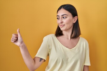 Hispanic girl wearing casual t shirt over yellow background looking proud, smiling doing thumbs up gesture to the side