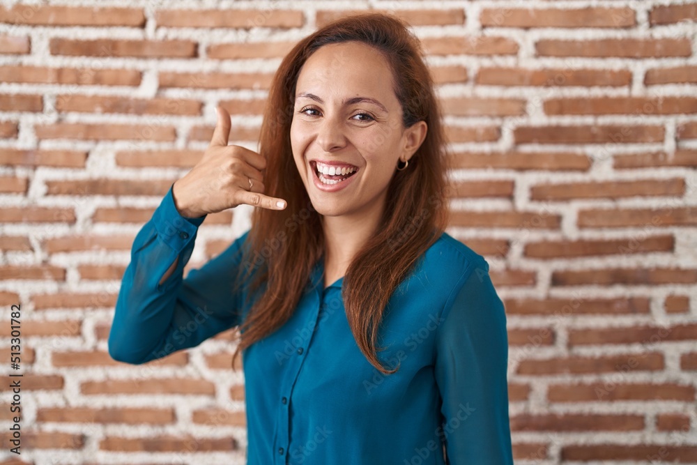 Sticker Brunette woman standing over bricks wall smiling doing phone gesture with hand and fingers like talking on the telephone. communicating concepts.