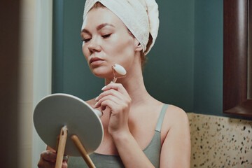 Young woman using jade facial roller for face massage sitting in bathroom, looking in the mirror
