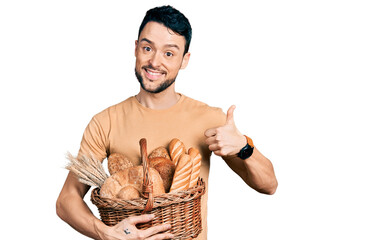 Hispanic man with beard holding wicker basket with bread smiling happy and positive, thumb up doing excellent and approval sign