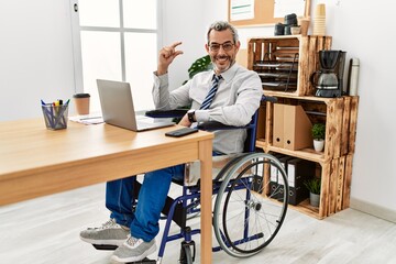 Middle age hispanic man working at the office sitting on wheelchair smiling and confident gesturing with hand doing small size sign with fingers looking and the camera. measure concept.