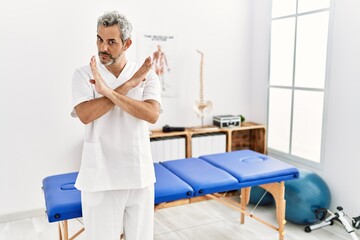 Obraz premium Middle age hispanic therapist man working at pain recovery clinic rejection expression crossing arms doing negative sign, angry face
