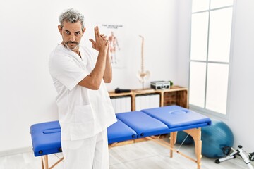 Middle age hispanic therapist man working at pain recovery clinic holding symbolic gun with hand gesture, playing killing shooting weapons, angry face