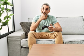 Fototapeta premium Middle age hispanic man playing video game sitting on the sofa serious face thinking about question with hand on chin, thoughtful about confusing idea