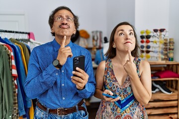 Middle age interracial couple at retail shop using smartphone thinking concentrated about doubt with finger on chin and looking up wondering