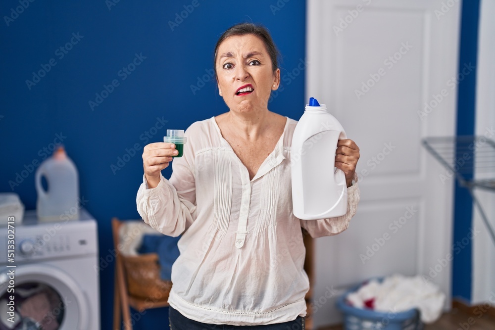 Sticker Middle age hispanic woman holding detergent bottle clueless and confused expression. doubt concept.