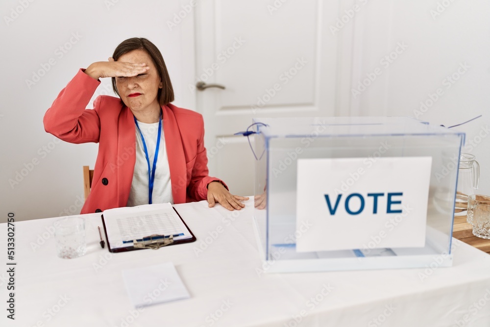 Sticker beautiful middle age hispanic woman at political election sitting by ballot covering eyes with hand,