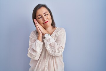 Middle age hispanic woman standing over blue background sleeping tired dreaming and posing with hands together while smiling with closed eyes.