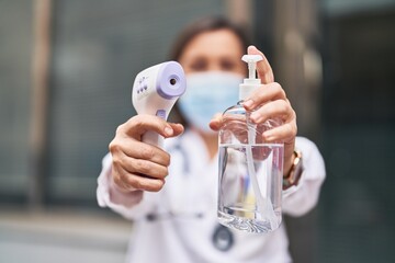 Middle age woman wearing doctor uniform and medical mask holding thermometer and sanitizer gel hands at street