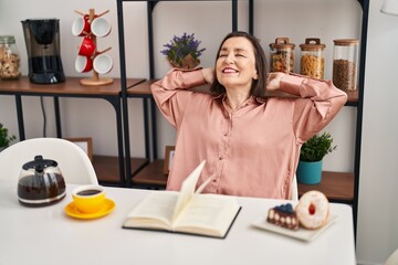 Middle age woman having breakfast relaxed with hands on head sitting on table at home