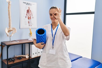 Young physiotherapist woman holding foam roll at the clinic smiling happy doing ok sign with hand on eye looking through fingers