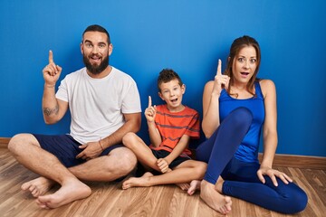 Family of three sitting on the floor at home pointing finger up with successful idea. exited and happy. number one.