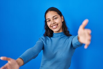 Fototapeta premium Young brazilian woman standing over blue isolated background looking at the camera smiling with open arms for hug. cheerful expression embracing happiness.