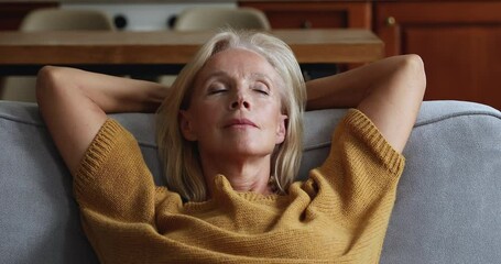 Peaceful calm mature elder lady sleeping with head resting on sofa back, relaxing with closed eyes, breathing fresh air, waking up, turning head, looking away, smiling - Powered by Adobe