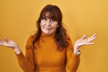 Middle age hispanic woman standing over yellow background clueless and confused expression with...