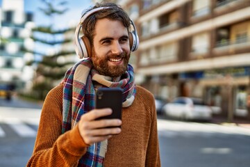 young caucasian man with beard listening to music with smartphone wearing headphones outdoors on a sunny day