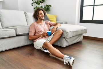 Young hispanic man drinking coffee sitting on the floor at home.