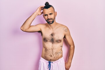 Fototapeta premium Young hispanic man shirtless wearing swimsuit confuse and wonder about question. uncertain with doubt, thinking with hand on head. pensive concept.